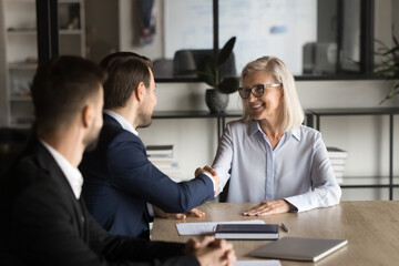 Positive confident younger and older business partners shaking hands on meeting, smiling, laughing, negotiating on contract, agreement, partnership. Senior team leader thanking employee with handshake