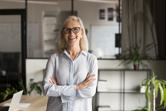 Cheerful confident old mature business woman in glasses posing in office with arms crossed, looking at camera, smiling, laughing, standing at workplace table. Professional head shot portrait