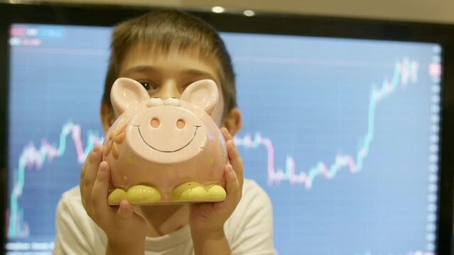 Early investment , financial education. Cute preschool child boy nocking a piggy bank in front of stock market chart. Wealth growth. High quality 4k footage