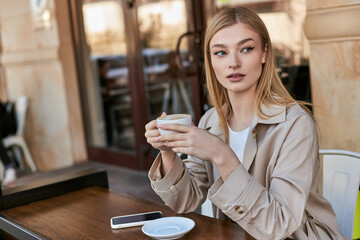 pretty young woman in trench coat enjoying cup of cappuccino while sitting near smartphone in cafe