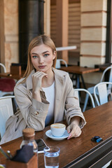 young blonde woman in trench coat enjoying her cup of cappuccino outdoors in cafe, weekend morning