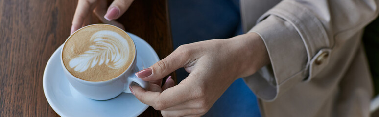 cropped young woman in trench coat holding cup with cappuccino in outdoor cafe, latte art banner