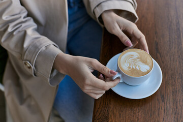 cropped view of young woman in trench coat holding cup with cappuccino in outdoor cafe, latte art