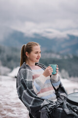 Female in a blanket in winter looking on a hill near a forest. Woman sitting and holding a cup and drinking hot coffee on winter holiday. Girl drink tea and relax on cabin terrace with a mountain view