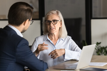 Serious elder professional woman talking to younger colleague man at workplace, working on project...