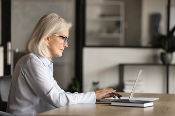 Serious blonde elder business woman in elegant glasses typing text on laptop, sitting at workplace table, using computer for online work communication, wireless Internet connection