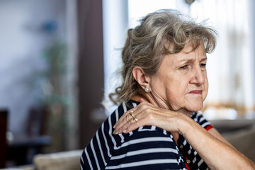 Portrait of senior woman suffering from neck pain while sitting on sofa at home - obrazy, fototapety, plakaty