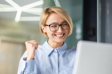 Successful woman celebrating victory and great achievement results at workplace, blonde business woman inside office received online notification on laptop, holding hand up triumph gesture.