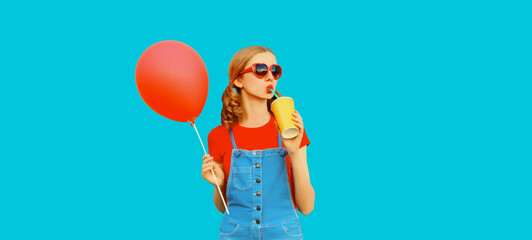Happy cheerful young woman drinks fresh juice with balloon looking away wearing red heart shaped sunglasses, jumpsuit on blue background