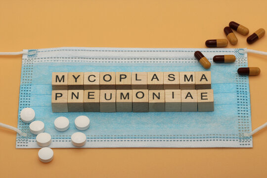 Mycoplasma pneumoniae. It is a human pathogen that causes the disease mycoplasma pneumonia. The words is laid out with wooden cubes on a surgical face mask. There are various pills lying around.