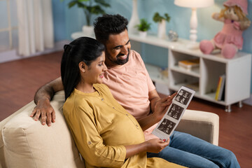 Happy Indian Pregnant women or wife with caring husband checking ultrasound scan reports at home -...