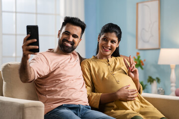 Happy Indian husband with pregnant wife taking selfie on mobile phone - concept of parenthood,...