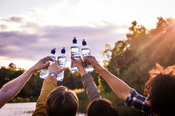 This image captures a group of friends raising water bottles towards the sky, reminiscent of a toast. The setting sun softly illuminates the scene, casting a warm glow and creating a radiant backdrop