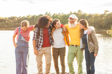 A diverse group of friends shares a moment of closeness by the lake, their body language suggesting comfort and familiarity with one another. The central figure stands out with a bright yellow shirt