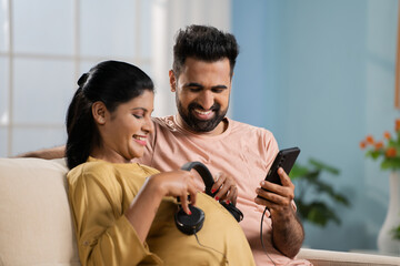 Happy indian husband with pregnant wife holding headphones on tummy by listening music her baby at home - concept of entertainment, maternity and relationship.
