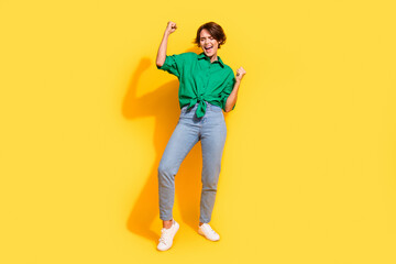 Full length photo of excited funky girl dressed green shirt rising fists shouting yeah isolated yellow color background