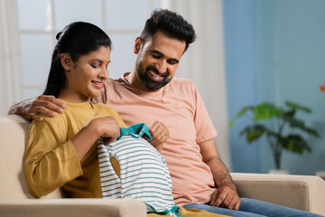 Happy Indian Pregnant woman checking baby dress by placing on tummy after given by husband -...