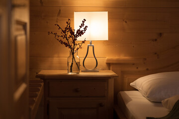 A cozy corner in a hotel room, on the bedside table there is a dimmed lamp and a bouquet of branches with red berries