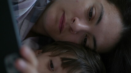 Close-up of mother and child looking at cellphone device screen laid in bed. Parent and son...