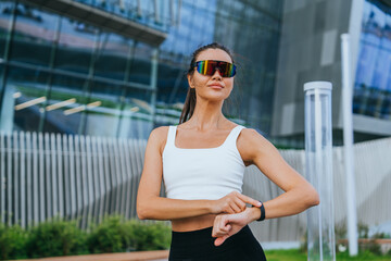 beautiful fit Asian woman in sunglasses, checks  pulse on fitness bracelet is standing in park, looking into distance against the background of a large glass building during break of running training.