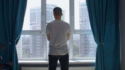 Young man standing near the window in a room with blue curtains. Media. Man waiting for something,...