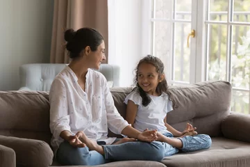 Foto op Plexiglas Young Indian woman her cute daughter folded fingers makes mudra gesture sit cross-legged on sofa laughing doing meditation practice. Parent teach to child yoga and good life habits, lifestyle concept © fizkes