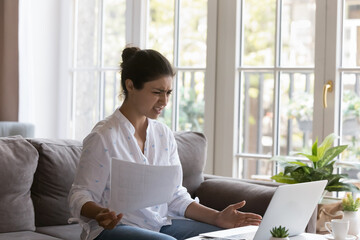 Indian woman holds papers staring at laptop screen feels annoyed looks angry, computer is stuck...