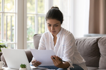 Young serious Indian woman housewife, entrepreneur sit on sofa holds paper document, learn agreement terms and conditions do paperwork at home looks attentive read formal important notice from bank - obrazy, fototapety, plakaty