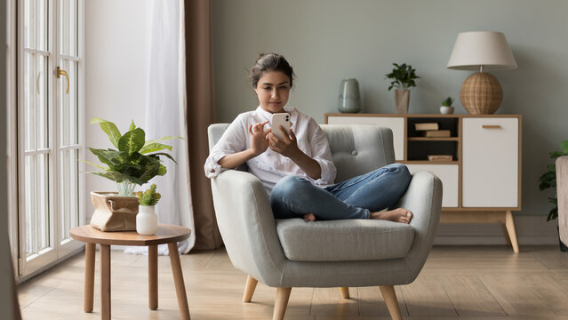 Attractive Indian woman relax on armchair in light fashionable living room holds smart phone chatting on-line, make order via internet electronic services. Leisure with tech, application usage concept