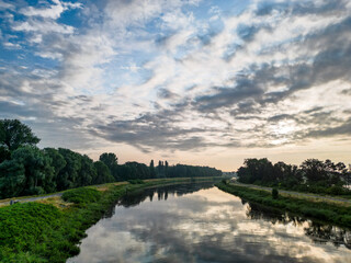 Fototapeta na wymiar This photograph captures the peaceful essence of a river landscape at dawn. The still waters create a perfect mirror, reflecting the awakening sky adorned with streaks of clouds. The lush riverbanks
