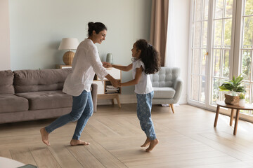 Indian mother and 6s daughter listen song hold hands dance barefoot on warm floor in cozy living room, spend carefree leisure move to favourite music. Happy homeowner family have fun at home concept