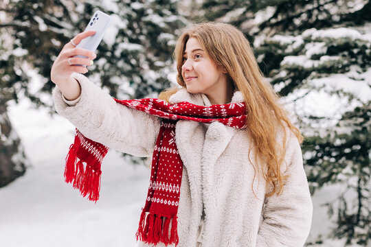 Young woman dressed in a fur coat and a red Christmas scarf takes a selfie outdoors in winter