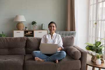 Happy Indian woman sit on sofa with laptop laughing staring at camera looks overjoyed enjoy modern technology, easy simple e-services and e-commerce retail services for comfort life. Freelance concept