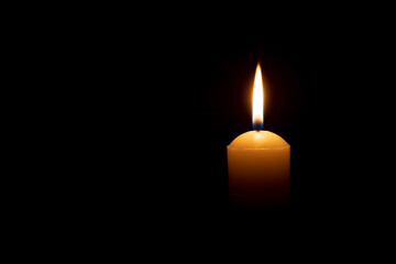 A single burning candle flame or light glowing on a big yellow candle on black or dark background on table in church for Christmas, funeral or memorial service with copy space - obrazy, fototapety, plakaty