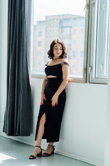Fototapeta premium Portrait of a young girl in a black dress standing by the window