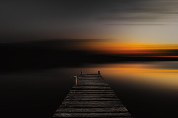 Old wooden boardwalk, dock in lake with abstract blurred background in dark evening. Abstract blurred sunset background