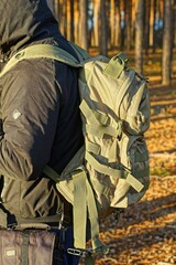 fashionable comfortable olive color military closed with plastic zippers vintage tourist backpack behind the back of a man dressed in a black jacket with a hood on his head in the forest 