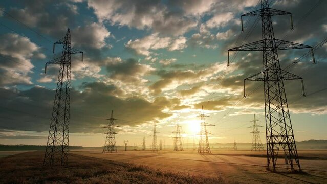 Flying towards electricity transmission towers in field at sunrise aerial drone shot. Sunlit high voltage poles and energy power lines with nature landscape and cloudscape