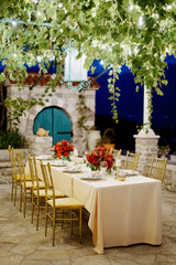 Set festive table in the courtyard of an ancient house with weaving green plants and glowing garlands