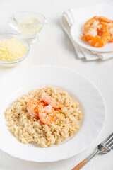 Portion of creamy homemade traditional italian risotto made of arborio rice cooked with broth decorated with shrimps or prawns and parmesan cheese served in plate with fork on white wooden table