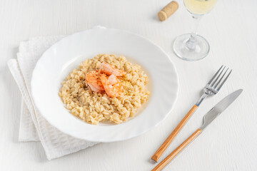 Creamy homemade traditional risotto made of fried and boiled arborio rice with shrimps seafood and parmesan cheese served in plate on napkin with tableware and glass of wine on white wooden table 