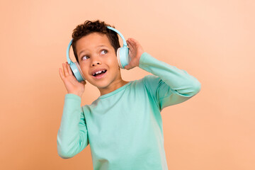 Photo of funky cheerful boy wear trendy turquoise clothes listen music wireless earbuds look empty space isolated on beige color background