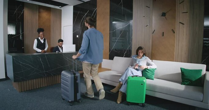 Handsome bearded man visiting hotel talking with qualified nice receptionist assisting clients with checking in and out. Good-looking young woman sitting on sofa in modern hotel.