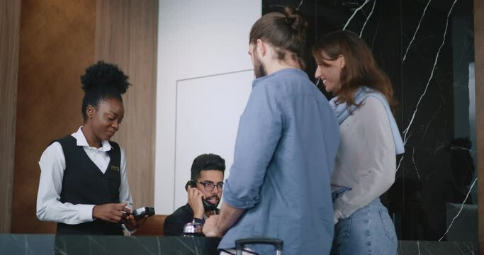 Back-view of cute young couple providing payment using credit card on reception with the help of polite and friendly African-American female receptionist. Luxury hotel concept.