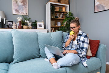 Young female freelancer in casual clothes sitting on comfortable couch and working remotely on...