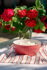 Cottage cheese homemade cheese in a red ceramic bowl, behind a bouquet of red roses, village atmosphere, sunny day, no people, close-up