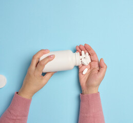 A woman's hand holding a plastic bottle and white oval pills on a blue background, top view.