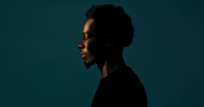 Profile of handsome attractive dark skinned male looking at light in darkstudio turning head to the camera looking wearing casual plain black t-shirt. Abstract futuristic concept.