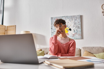 young transgender freelancer in pink sweater holding pen and looking at laptop on desk, remote work