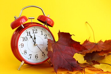 There is a red alarm clock with yellow autumn leaves on a yellow background.